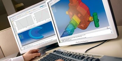 An engineer analyzes a 3D simulation of a mechanical component on dual monitors, displaying a finite element analysis (FEA) model with color-coded stress distribution.