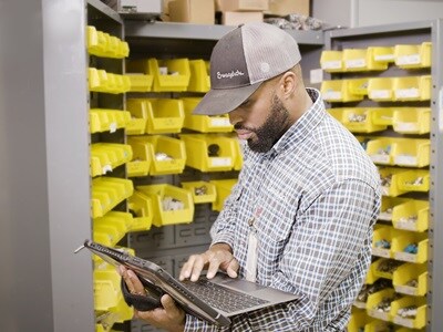 A warehouse worker wearing a Swagelok cap and a plaid shirt uses a tablet to manage inventory in a storage room filled with yellow bins containing small parts.